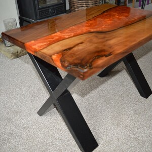 May include: A wooden coffee table with a live edge top and a black metal X-shaped base. The top is finished with a clear epoxy resin that has a red and orange swirl.