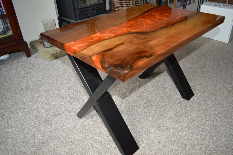 May include: A wooden coffee table with a live edge top and a black metal X-shaped base. The top is finished with a clear epoxy resin that has a red and orange swirl.