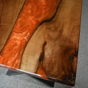 May include: A close-up of a wooden table top with a large orange resin river running through the center. The wood is a dark brown color and the resin is a bright orange.
