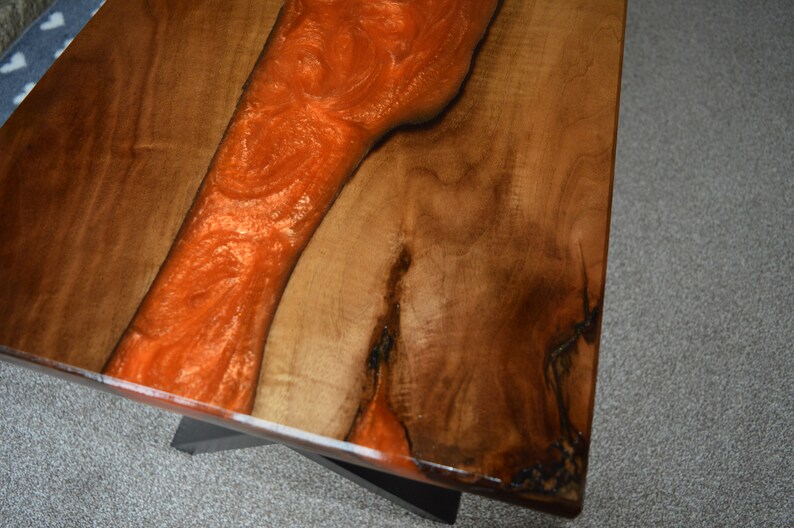 May include: A close-up of a wooden table top with a large orange resin river running through the center. The wood is a dark brown color and the resin is a bright orange.