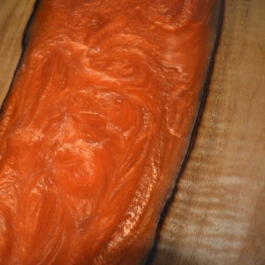 May include: A close-up of a wooden table top with a large orange resin inlay. The resin has a swirling pattern and a glossy finish.