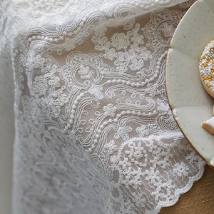 May include: White lace fabric with a delicate floral pattern. The lace has a scalloped edge and is shown on a brown surface.