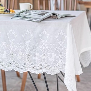 May include: A white tablecloth featuring detailed embroidered floral and geometric designs along the border. The cloth is laid over a wooden table, offering a clean, elegant aesthetic. A magazine and a plate of food are on the table.
