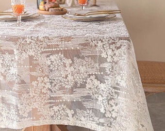 May include: White lace tablecloth with a floral pattern. The tablecloth is draped over a table with a wooden surface.