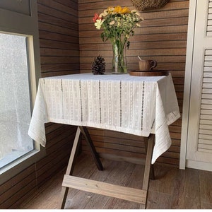 May include: White cotton tablecloth with a delicate lace pattern and vertical stripes. The tablecloth is draped over a wooden table with a rustic X-shaped base.