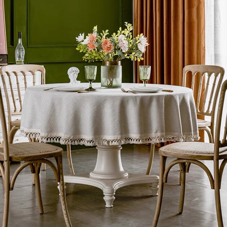 May include: A round white table with a beige linen tablecloth with a white fringe trim. The table is set with four chairs and a vase of flowers in the center.
