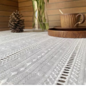May include: White embroidered tablecloth with a delicate pattern. The tablecloth is laid on a wooden table with a wooden tray and a brown cup.