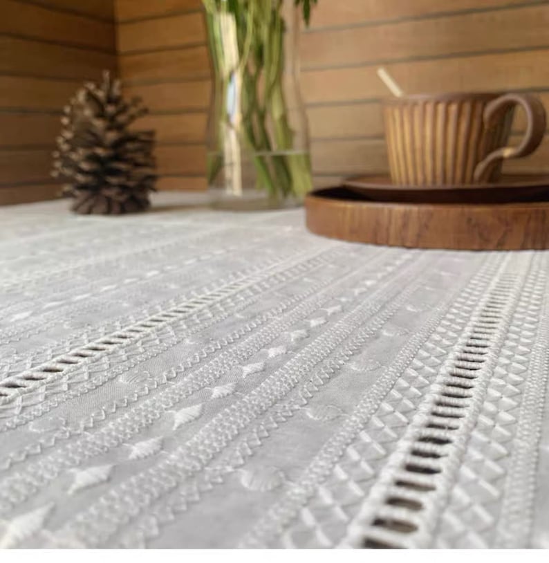 May include: White embroidered tablecloth with a delicate pattern. The tablecloth is laid on a wooden table with a wooden tray and a brown cup.