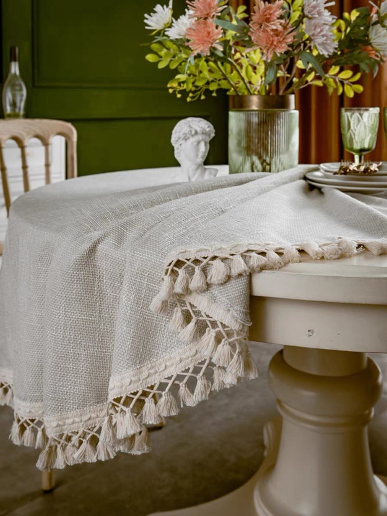 May include: A round, off-white tablecloth with a fringe border. The tablecloth is made of a woven fabric and has a subtle texture. The tablecloth is draped over a round table with a white pedestal base.
