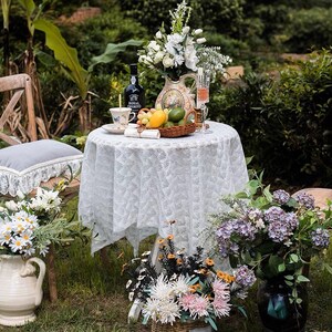 May include: A white lace tablecloth with a floral pattern covers a round table set for a garden party. A bottle of wine, a glass of wine, a plate of fruit, and a vase of white flowers are on the table.
