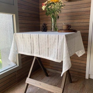 May include: White cotton tablecloth with a delicate embroidered pattern of small dots and vertical lines. The tablecloth is draped over a wooden table with a rustic finish.