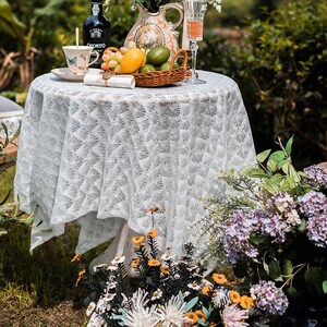 May include: White lace tablecloth with a delicate floral pattern, set on a round table with a white base. The table is decorated with a pitcher, a glass of wine, a plate with a cup and saucer, a bowl of fruit, and a basket of limes. The table is set in a garden setting with green grass and flowers.