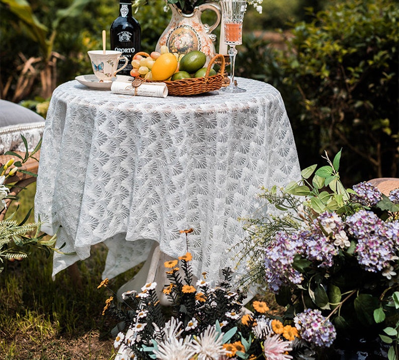 May include: White lace tablecloth with a delicate floral pattern, set on a round table with a white base. The table is decorated with a pitcher, a glass of wine, a plate with a cup and saucer, a bowl of fruit, and a basket of limes. The table is set in a garden setting with green grass and flowers.