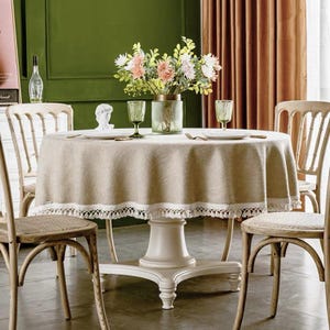 May include: A round, beige linen tablecloth with white lace trim and tassels hangs over a white pedestal table. The table is set with two chairs and a vase of pink and white flowers.