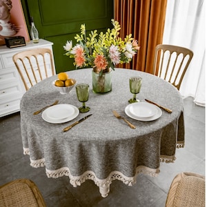 May include: A round gray tablecloth with a white fringe border. The tablecloth is set with two place settings, each with a white plate, gold silverware, and a clear glass. A vase with pink and yellow flowers sits in the center of the table.
