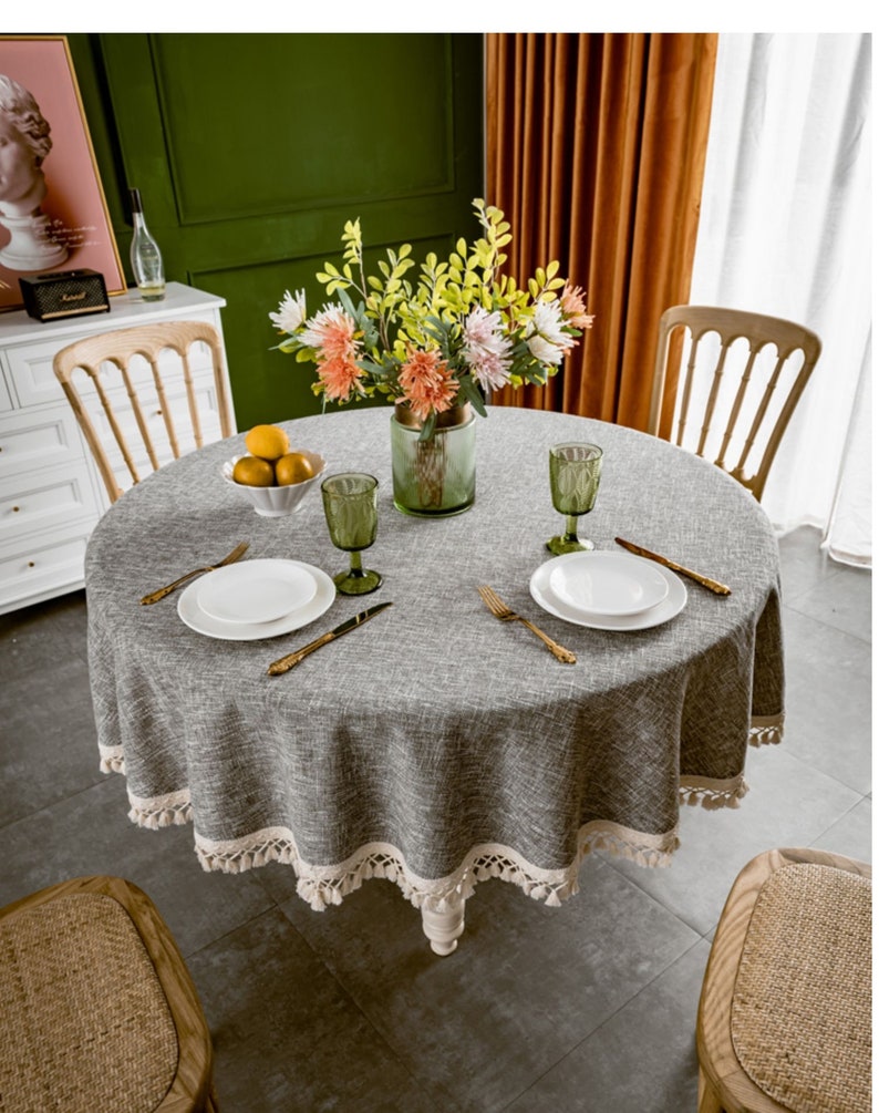 May include: A round gray tablecloth with a white fringe border. The tablecloth is set with two place settings, each with a white plate, gold silverware, and a clear glass. A vase with pink and yellow flowers sits in the center of the table.