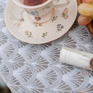 May include: A white lace tablecloth with a delicate, repeating pattern of small circles and curved lines. A white teacup and saucer with floral and butterfly designs are on top of the tablecloth.