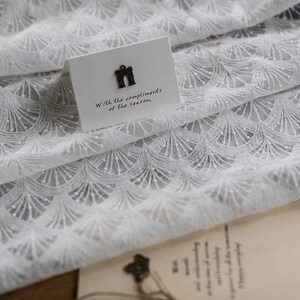 May include: White lace fabric with a delicate, repeating pattern of small arches. A small card with the text "With the compliments of the season." is placed on top of the fabric.