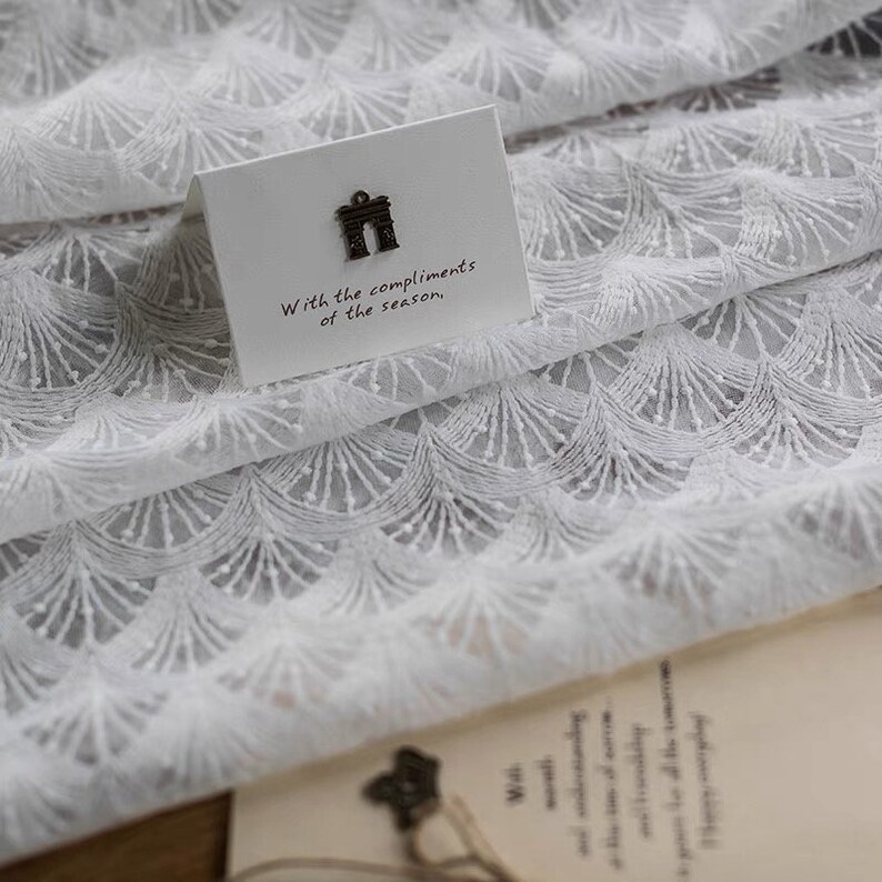 May include: White lace fabric with a delicate, repeating pattern of small arches. A small card with the text "With the compliments of the season." is placed on top of the fabric.