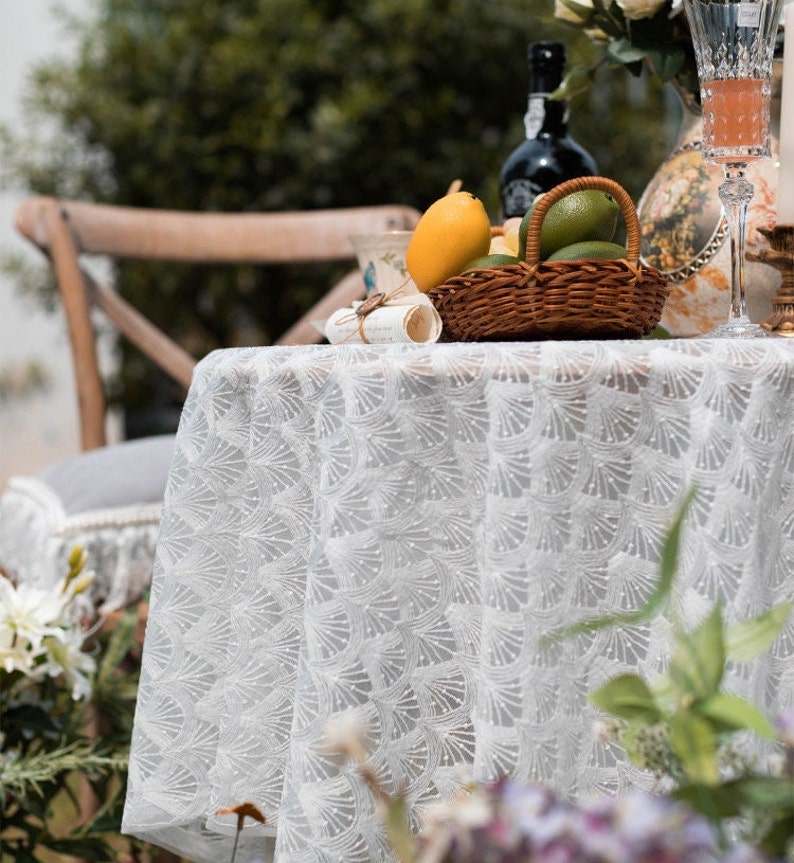 May include: White lace tablecloth with a delicate pattern, set for a summer picnic with a basket of lemons and limes, a glass of wine, and a vintage teacup.