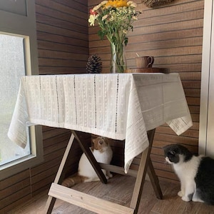 May include: White lace tablecloth with a delicate pattern, draped over a wooden table with two cats sitting underneath. The table is set with a cup and saucer.