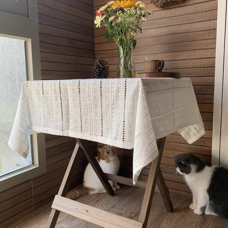 May include: White lace tablecloth with a delicate pattern, draped over a wooden table with two cats sitting underneath. The table is set with a cup and saucer.