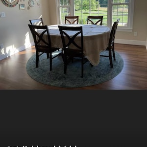 May include: A round dining table with a beige tablecloth and four chairs. The table is set with placemats and silverware. The chairs are brown wood with a cross-back design. The table is in a room with a large window overlooking a green lawn.