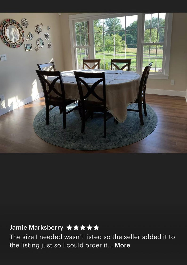 May include: A round dining table with a beige tablecloth and four chairs. The table is set with placemats and silverware. The chairs are brown wood with a cross-back design. The table is in a room with a large window overlooking a green lawn.