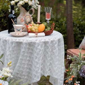 May include: A white lace tablecloth with a delicate floral pattern covers a round table. A white teacup with a butterfly design sits on a saucer next to a bottle of wine. A wicker basket filled with fruit, including grapes, a mango, and limes, is on the table. A white candle and a glass of pink liquid are also on the table.