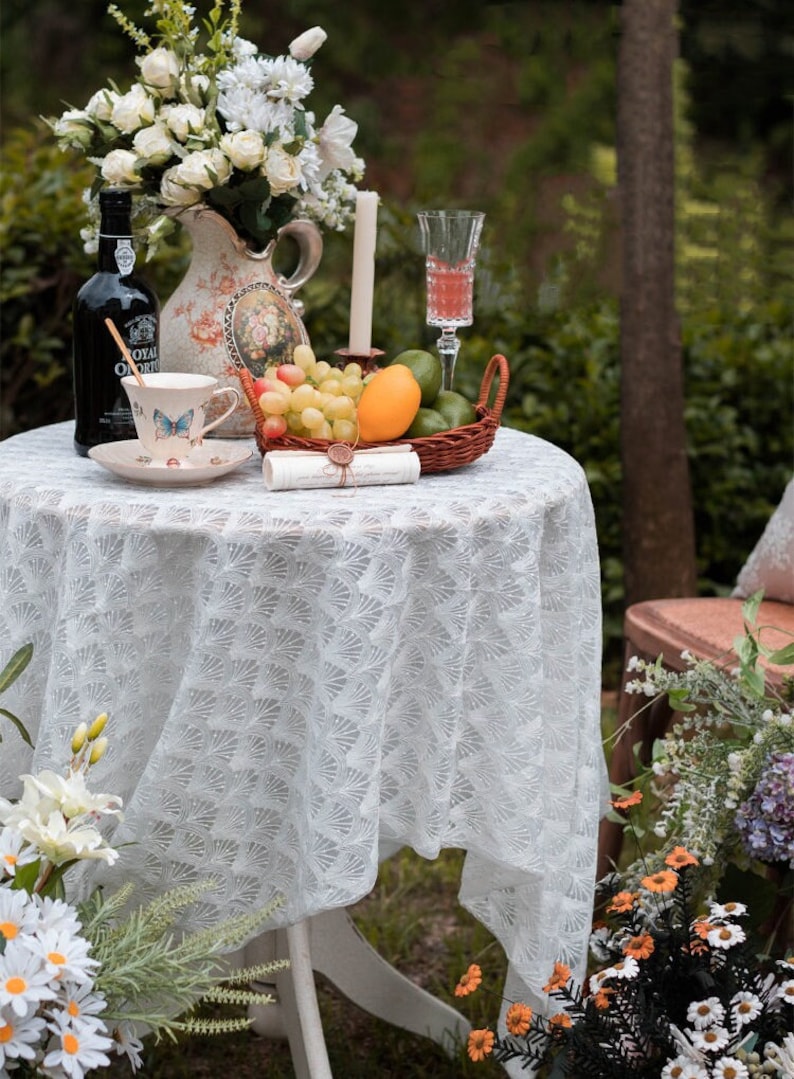 May include: A white lace tablecloth with a delicate floral pattern covers a round table. A white teacup with a butterfly design sits on a saucer next to a bottle of wine. A wicker basket filled with fruit, including grapes, a mango, and limes, is on the table. A white candle and a glass of pink liquid are also on the table.