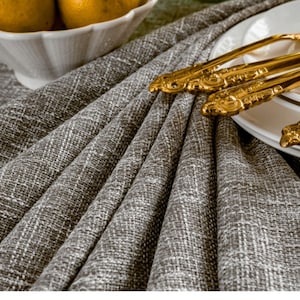 May include: A close-up of a gray linen tablecloth with a set of gold-plated silverware on a white plate.