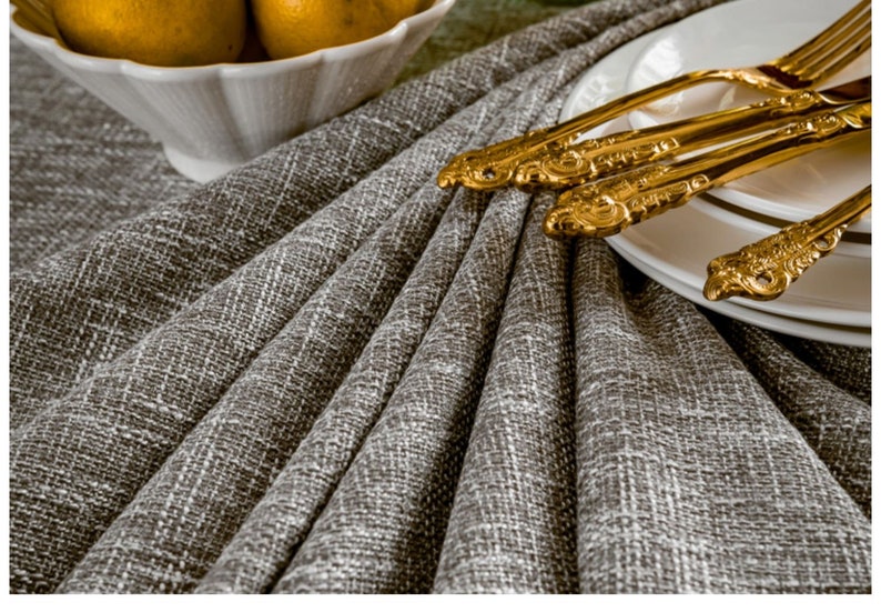 May include: A close-up of a gray linen tablecloth with a set of gold-plated silverware on a white plate.