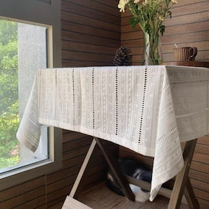May include: White crochet tablecloth with a delicate pattern, draped over a wooden table with a vase of flowers and a cup on a side table.