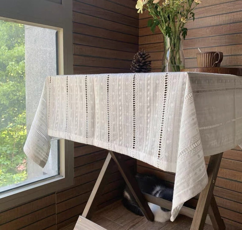 May include: White crochet tablecloth with a delicate pattern, draped over a wooden table with a vase of flowers and a cup on a side table.