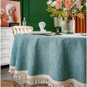 May include: A teal blue round tablecloth with a white crocheted trim and fringe. The tablecloth is on a white table with a wooden chair and a vase of flowers.