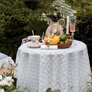 May include: A white lace tablecloth with a delicate floral pattern covers a round table. A vintage pitcher with floral designs holds a bouquet of white flowers. A teacup with a butterfly design sits on a saucer next to a basket of fruit, including grapes, lemons, and limes. A candle and a glass of wine complete the scene.