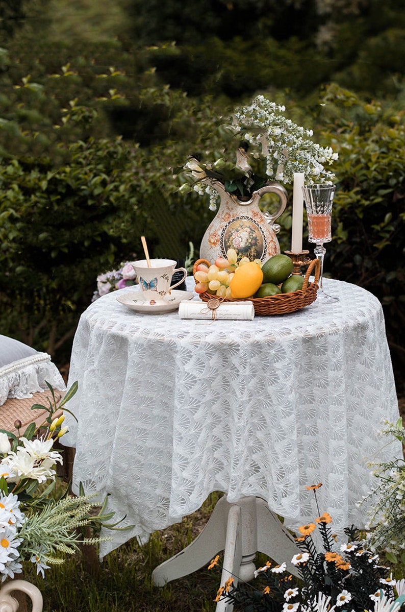 May include: A white lace tablecloth with a delicate floral pattern covers a round table. A vintage pitcher with floral designs holds a bouquet of white flowers. A teacup with a butterfly design sits on a saucer next to a basket of fruit, including grapes, lemons, and limes. A candle and a glass of wine complete the scene.