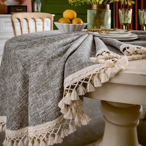 May include: A round gray tablecloth with a white crocheted trim and tassels. The tablecloth is on a white table with a pedestal base.