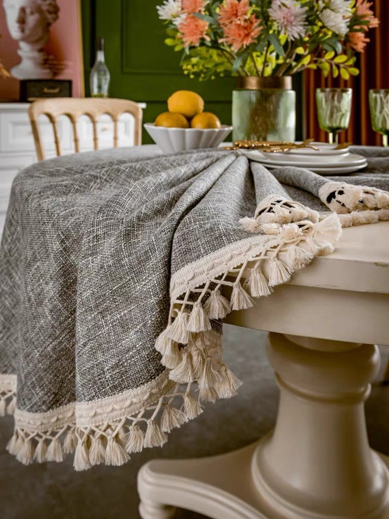 May include: A round gray tablecloth with a white crocheted trim and tassels. The tablecloth is on a white table with a pedestal base.