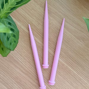 May include: Three light pink knitting needle point protectors. The protectors are long and tapered, with a small ring at the base. The text "Heavenly Knits Handmade with love" is at the bottom of the image.