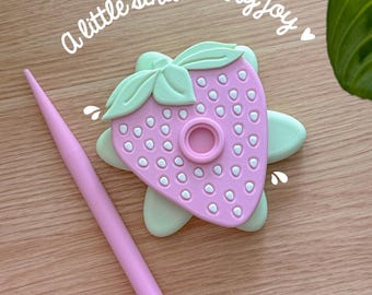 Peut inclure: Un objet rose en forme de fraise avec un design de feuilles vert clair et un stylet rose. Le texte "a little strawberry joy" est écrit en cursive. Les mots "Heavenly Knits Handmade with love" sont également visibles.