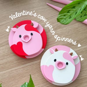 以下が含まれることがあります： バレンタインデーの牛の糸スピナーが2つ。1つは赤とピンク、もう1つは白とピンクです。スピナーは、ピンクの鼻と茶色の角を持つ牛の顔のデザインです。「Valentine's cow yarn spinners」というテキストが画像にあります。