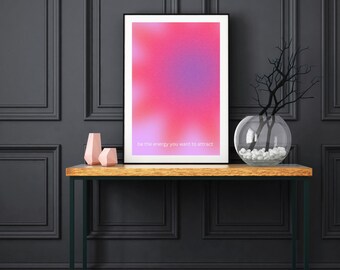 May include: A black framed poster with a pink and purple gradient background and the text "be the energy you want to attract". The poster is on a wooden console table with two pink vases and a glass vase with white stones.