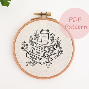 以下が含まれることがあります： 「Coffee, Read, Repeat」の文字とコーヒーカップが描かれた本の山をあしらった刺繍アート。周りには花のモチーフが施されています。木製のフレームに、淡いベージュ色の布が張られています。