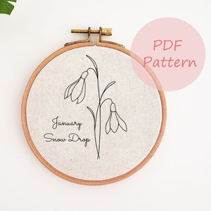 Op de afbeelding: Een witte borduurring met een bruin houten frame. De ring heeft een zwarte lijntekening van een sneeuwklokje met de tekst "January Snow Drop".