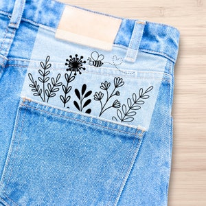 Puede incluir: Un bolsillo de mezclilla azul con una hoja de plástico transparente en su interior. La hoja tiene un diseño floral en blanco y negro con una abeja volando sobre las flores.