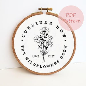May include: A wooden embroidery hoop with a white fabric center featuring a black floral design and the text "CONSIDER HOW THE WILDFLOWERS GROW" with "LUKE 12:27" below. The hoop has a gold-colored clasp and the words "PDF Pattern" in a pink circle.