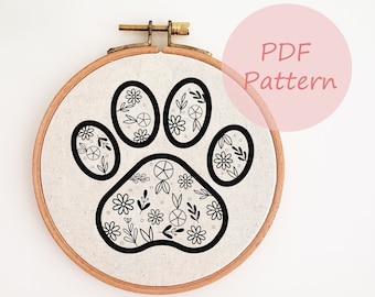 Könnte beinhalten: Ein schwarz-weißes Stickmuster einer Pfote mit floralen Details. Das Muster ist auf einem runden Stück Stoff in einem hölzernen Stickrahmen dargestellt. Der Text "PDF Pattern" ist auf einem rosa Kreis dargestellt.