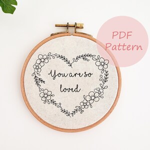May include: A round embroidery hoop with a linen fabric featuring a heart-shaped floral design and the words "You are so loved" in black script. The hoop is made of wood and has a brass clasp.