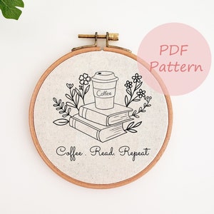 以下が含まれることがあります： リネンの生地に、コーヒーカップ、本、花の要素を黒い線で描いた刺繍枠。イラストの下には「Coffee. Read. Repeat」の文字。ピンクの円の中に「PDF Pattern」の文字があります。